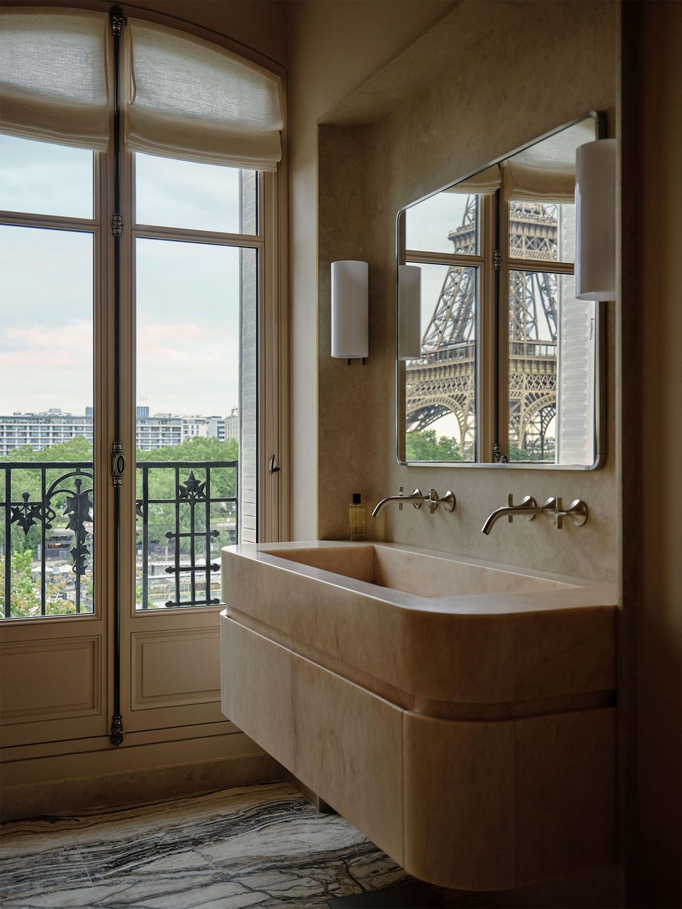 bathroom with a large light rose marble sink with two sets of silver matte fittings, sconces flank a square mirror that reflects the eiffel tower, textured walls, patterned rug, windowed door opens to wrought iron balcony