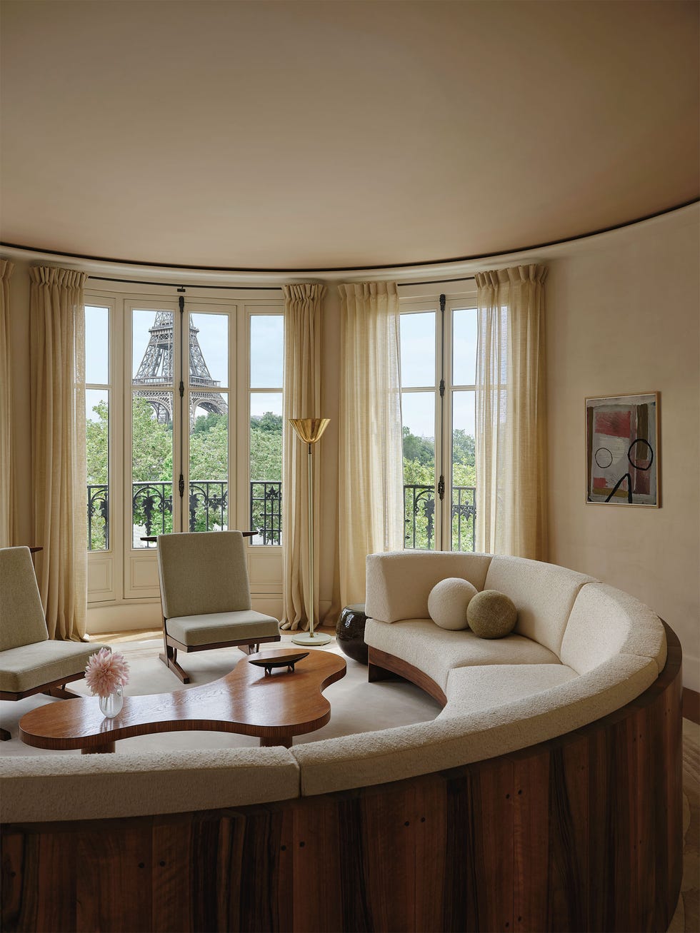 a curved living room has a large circular wood frame sofa with light colored cushions and ball shaped accent pillows, an amorphous wood cocktail table, two armless chairs with cushions, windows with a view of eiffel tower