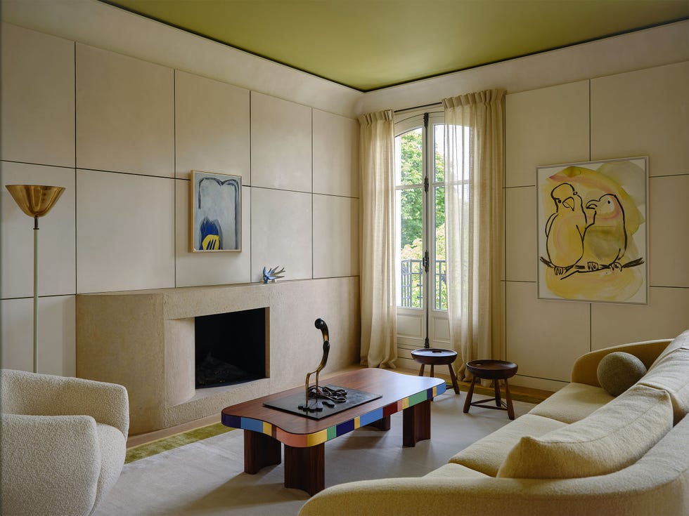 den has rectangular paneled walls, torchiere, boucle chair, curved fireplace, cocktail table with curved edge in bright colors, metal sculpture on table, curved back sofa, light rug, window, chartreuse ceiling, two artworks