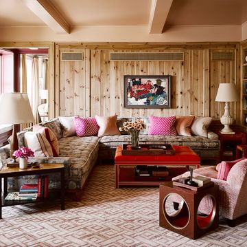 room in knotty pine and pink geometric carpeting and an l shaped sofa with tufted seats and multiple pillows in varying shades and designs of pink and a square red coffee table and a matching club chair in a dusty pink and white lamps