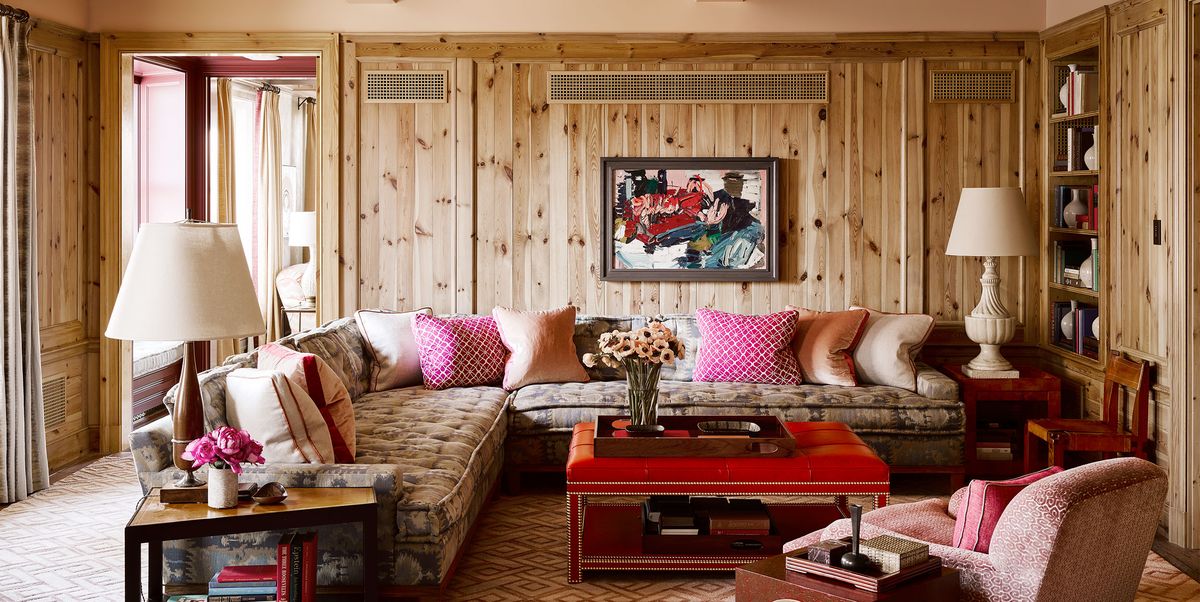 room in knotty pine and pink geometric carpeting and an l shaped sofa with tufted seats and multiple pillows in varying shades and designs of pink and a square red coffee table and a matching club chair in a dusty pink and white lamps