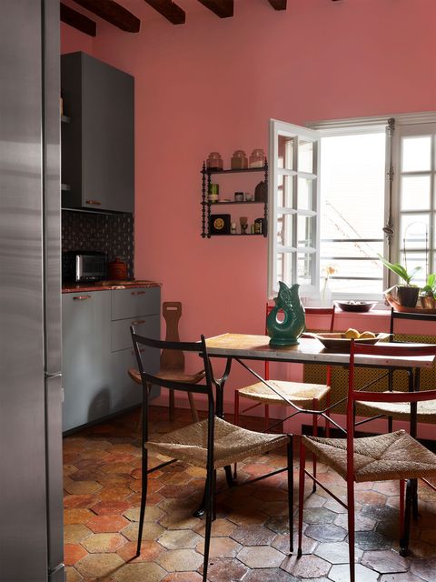 dining area in  kitchen with terracotta multihued hexagonal tiles and a salmon painted wall and a thin plank table at center with rush seats and on the back wall is an open window