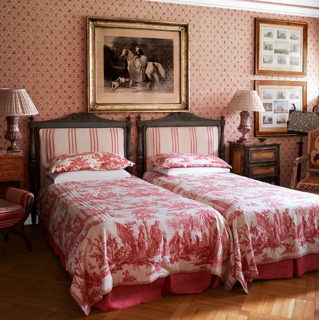 room with close twin beds with reddish pink toile covers and complementary striped fabric headboards with pleated shades on the nightstand lamps and patterned wallpaper in a similar hue