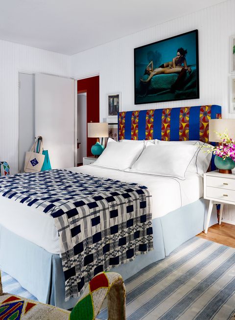 white room with white sheets and a rustic navy blue and white cover at the foot of the bed with a bright blue headboard with embroidered stripes of large red and yellow flowers and a chambray blue and white linen rug on a wooden floor
