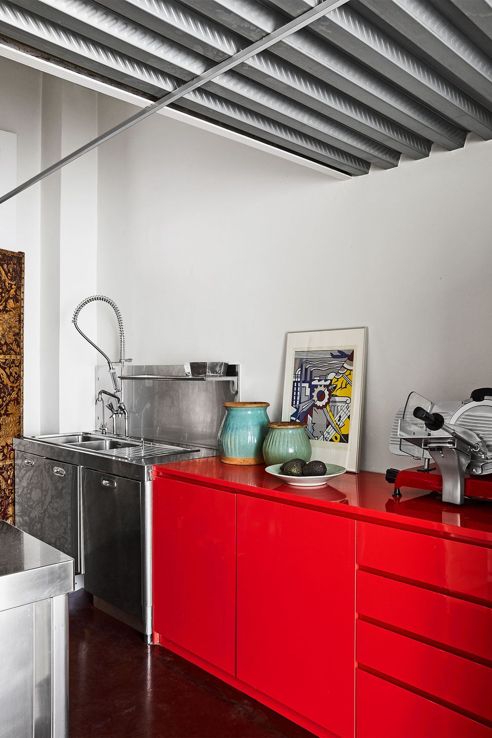 detail of a kitchen with long red lacquer counter height cabinet with with a meat slicer on it and several terra cotta jars and a piece of artwork standing against the wall and there is a a stainless sink and cabinet next to it with a long springy faucet