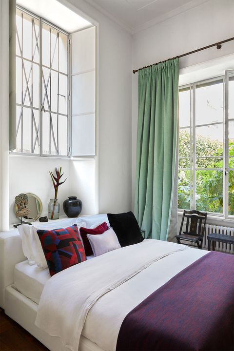 simple white bedroom with a white bed with a wide windows with celadon curtains and a dark eggplant colored bedspread on it and somewhat high above the bed is a white window with decorative protective bars