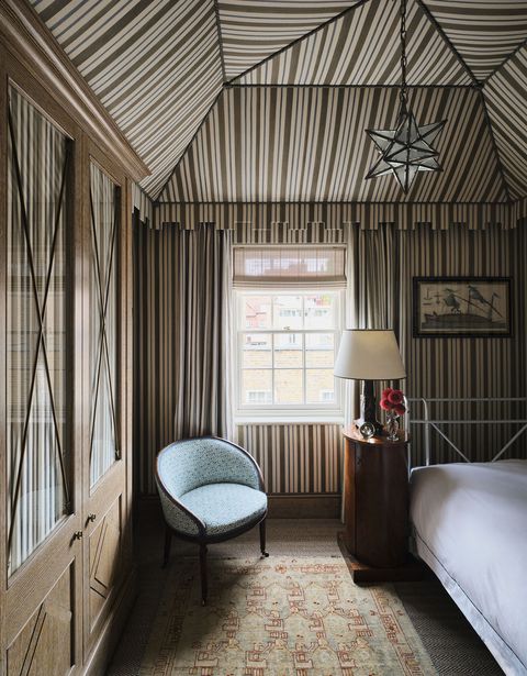 a tented dressing room has brown and white striped fabric walls and ceiling, built in closets, a blue fabric mahogany tub chair, a lamp on a small wood cabinet, a daybed, a patterned rug, and a star pendant