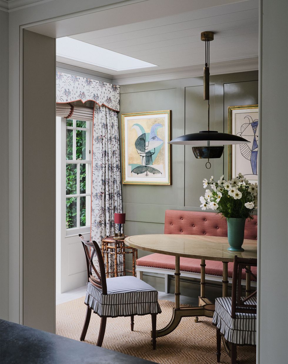 in a dining area are wood chairs with skirted fabric seats, an oval wood table, an upholstered banquette against a sage colored wall, a hexagonal side table, floral curtains, and pochoir artworks