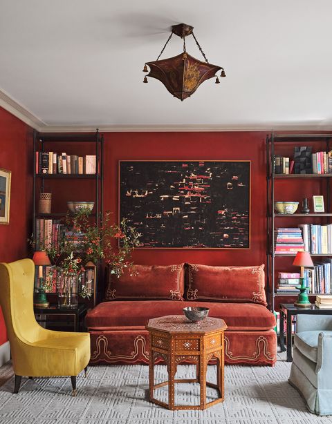 a deep red drawing room has a high back yellow chair, a velvet banquette with decorative cord work flanked by shelves of books and curios, a light blue upholstered chair, and an octagonal cocktail table