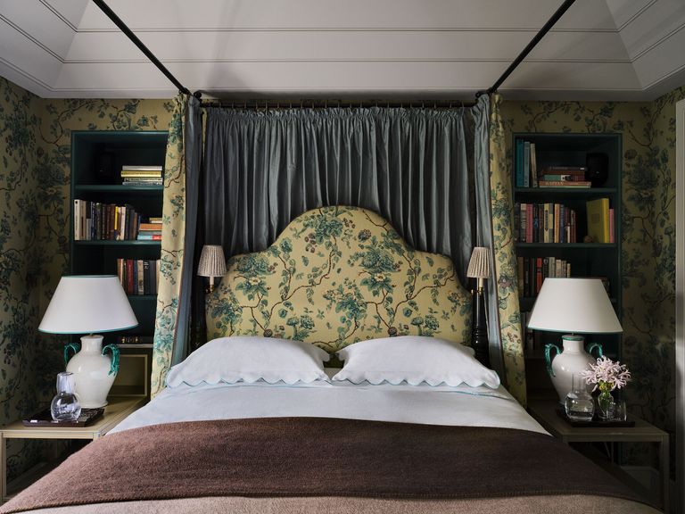 walls, bed curtains and headboard are in a fabric with a green floral pattern on a light yellow background, sconces are attached to headboard, side tables with lamps, and bed is flanked by bookshelves