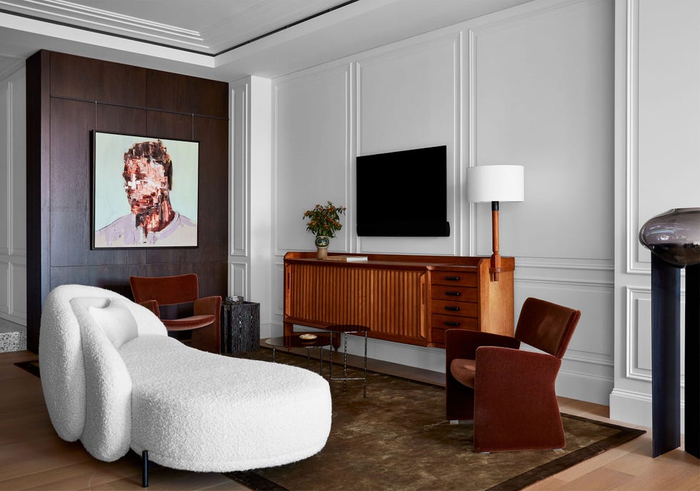 white paneled room with one dark wall on the left with a portrait hanging on it and a dark console against the white wall and a curvy armless chaise like sofa on which to recline and watch the tv hanging over the console