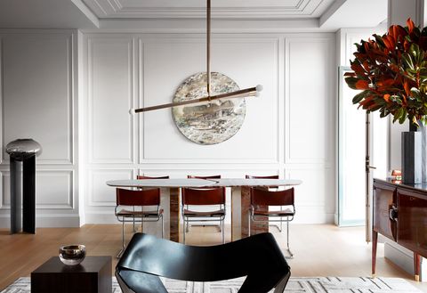 dining area with a skinny pedestal table and simple chairs in a large space with a white paneled wall in the background on which a round piece of art hangs and in the foreground is a wavy black chair that forms part of the living room