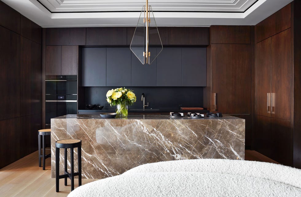 a dark panelled kitchen with a light brown and white splashed marble island and two dark wood stools with light tops and a white rounded table edge in the foreground