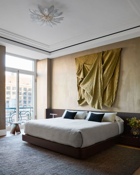 bed on a dark base with oatmeal colored bedspreads and a similar colored rug and above the bed is a yellow textile draped like an artwork and on the left are open glass doors through which you can see a small balcony