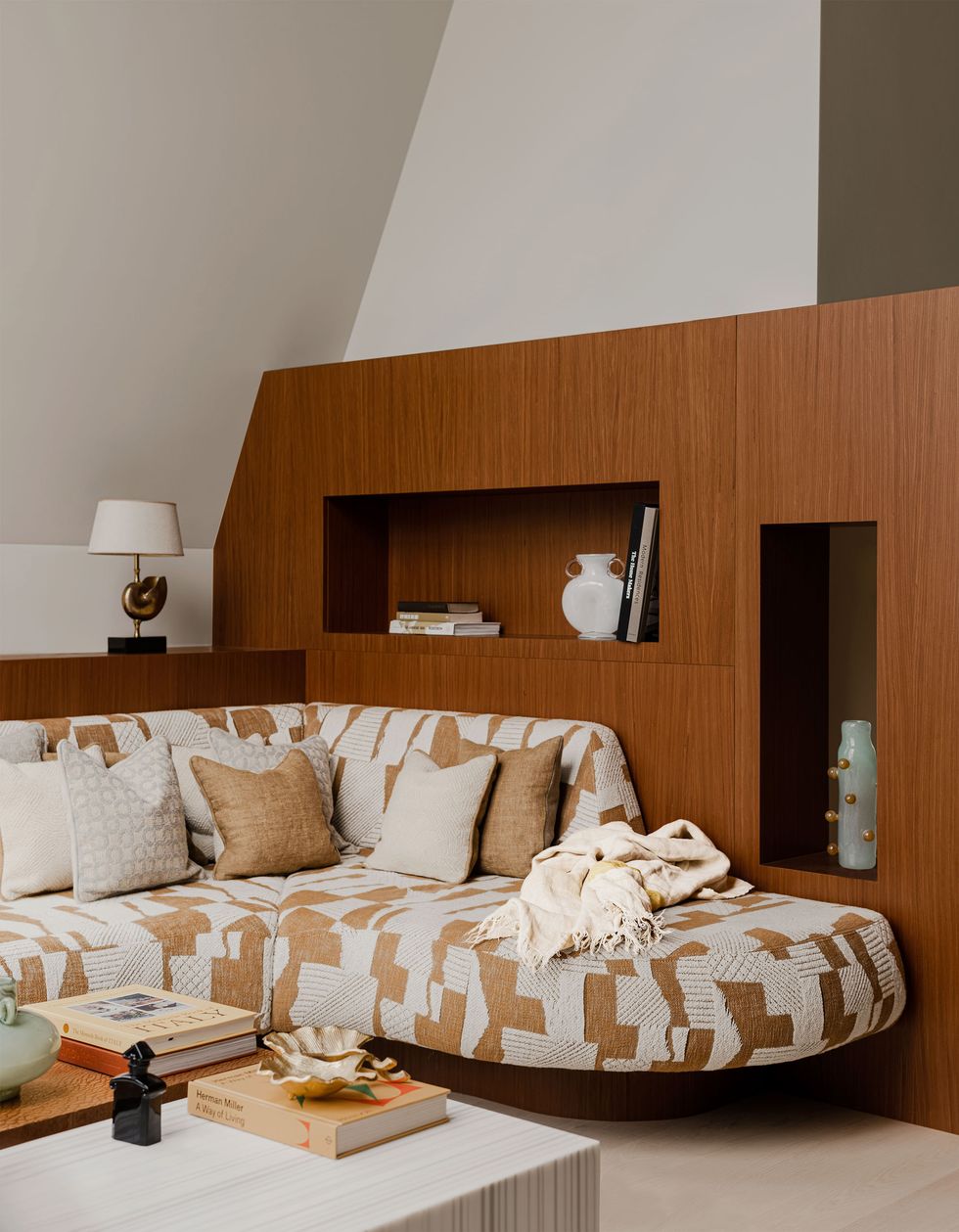 beige and white geometric design rounded sofa up against a dark honey color wall unit with a few cutout shelves and in the foreground are two low tables one white striated and other other textured wood with various objects and books on them