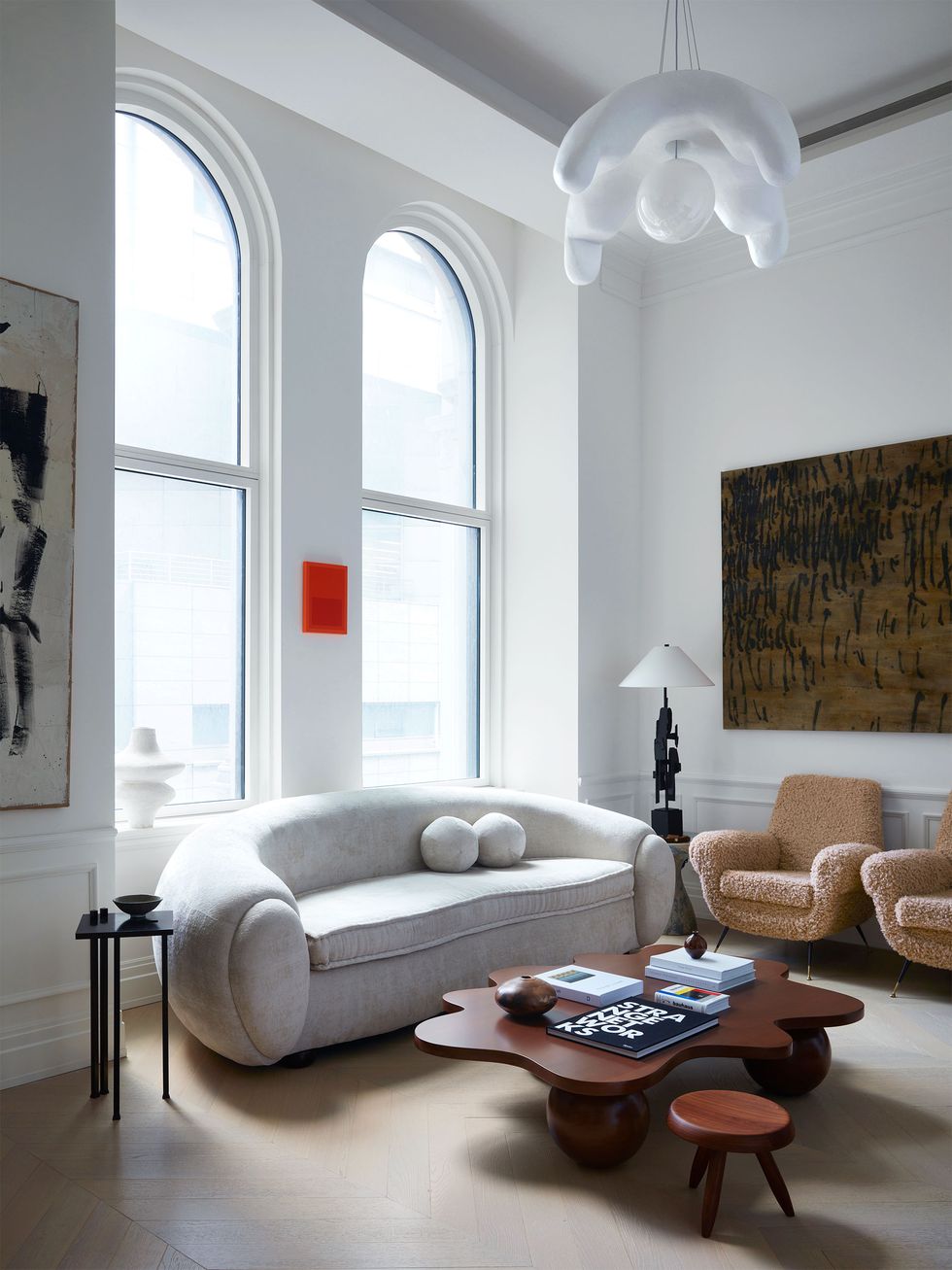 a living room has white glass pendant, tall arched windows with a curved sofa in front and side tables and a lamp, a wavy wood cocktail table and three legged stool, two tan boucle chairs, and two large artworks