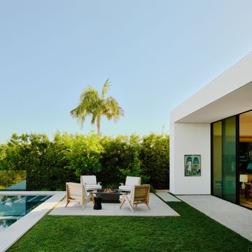 outside the living room, with its glass doors and white concrete frame, is a long rectangular infinity pool, a firepit and four wood chairs with cushions next to a row of shrubs and the los angeles cityscape beyond