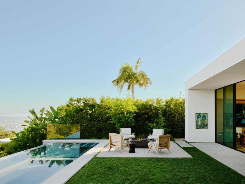 outside the living room, with its glass doors and white concrete frame, is a long rectangular infinity pool, a firepit and four wood chairs with cushions next to a row of shrubs and the los angeles cityscape beyond