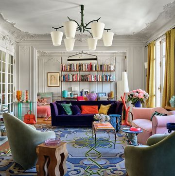 a living room with a blue rug and a book shelf and a purple sofa at center, a pink loveseat on the side, and a six cup chandelier at center with very french frosty rococo molding all around