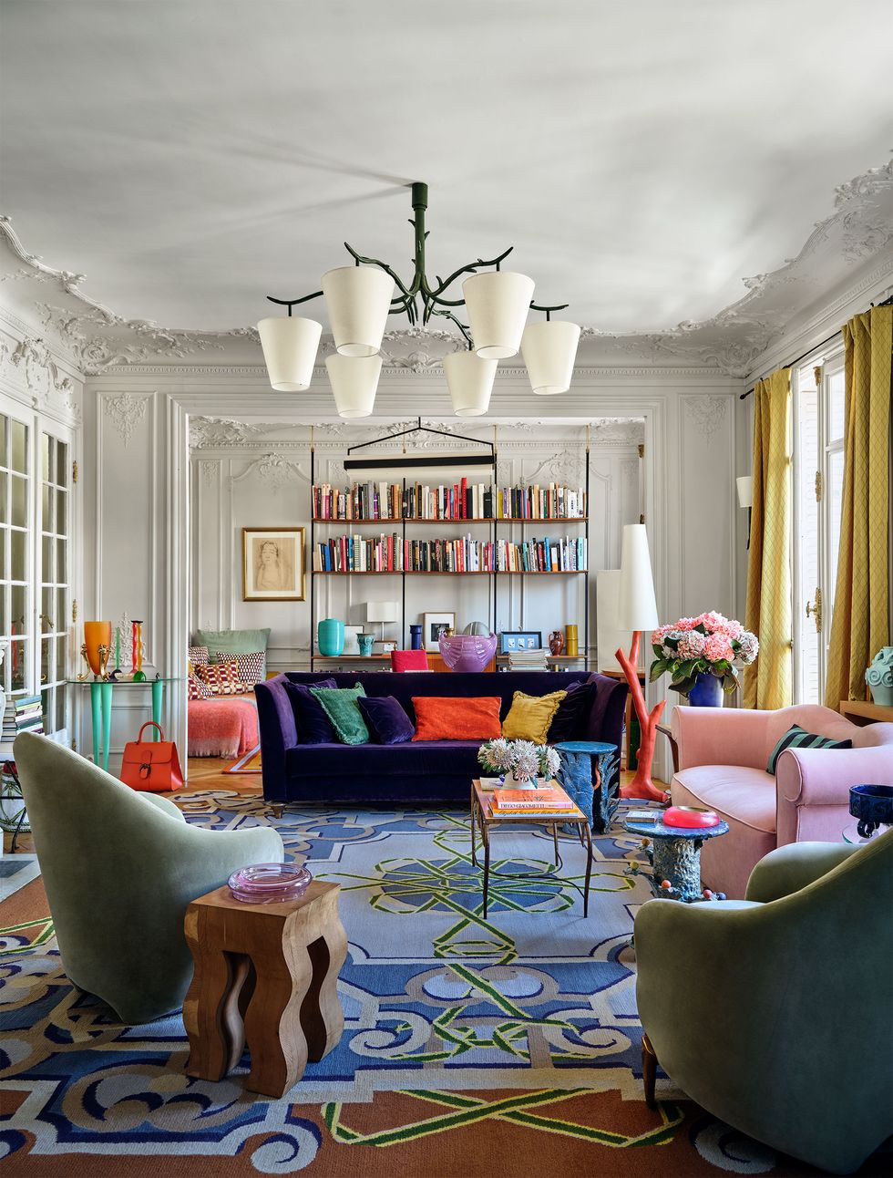 a salon has a six cup chandelier in center, bookshelves in background, a purple and a pink sofa, two upholstered armchairs, a floor lamp with branch shaped vase, several side tables, blue patterned rug