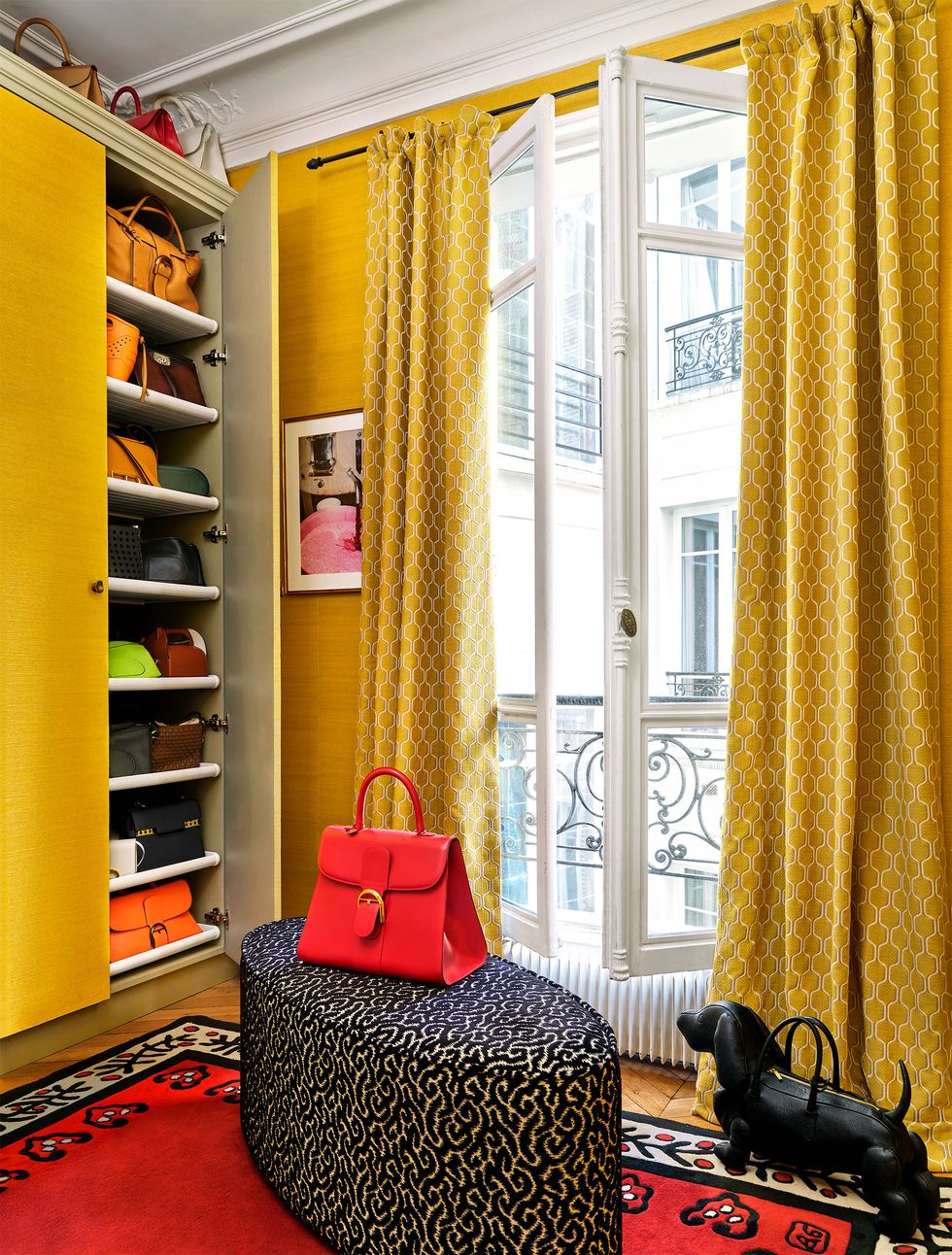 in a yellow dressing room is a large window with yellow curtains, an armoire filled with tens of leather handbags on shelves, patterned oval ottoman with a red bag atop, dog shaped handbag on floor, red rug