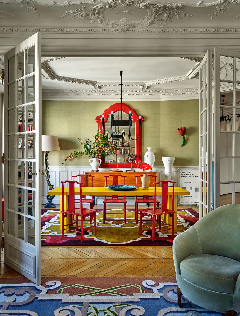 french doors open to a dining room with a bright yellow table and five red chairs, a sideboard with vases, a large red framed mirror, red flower sconce, floor lamp, burgundy and green patterned rug