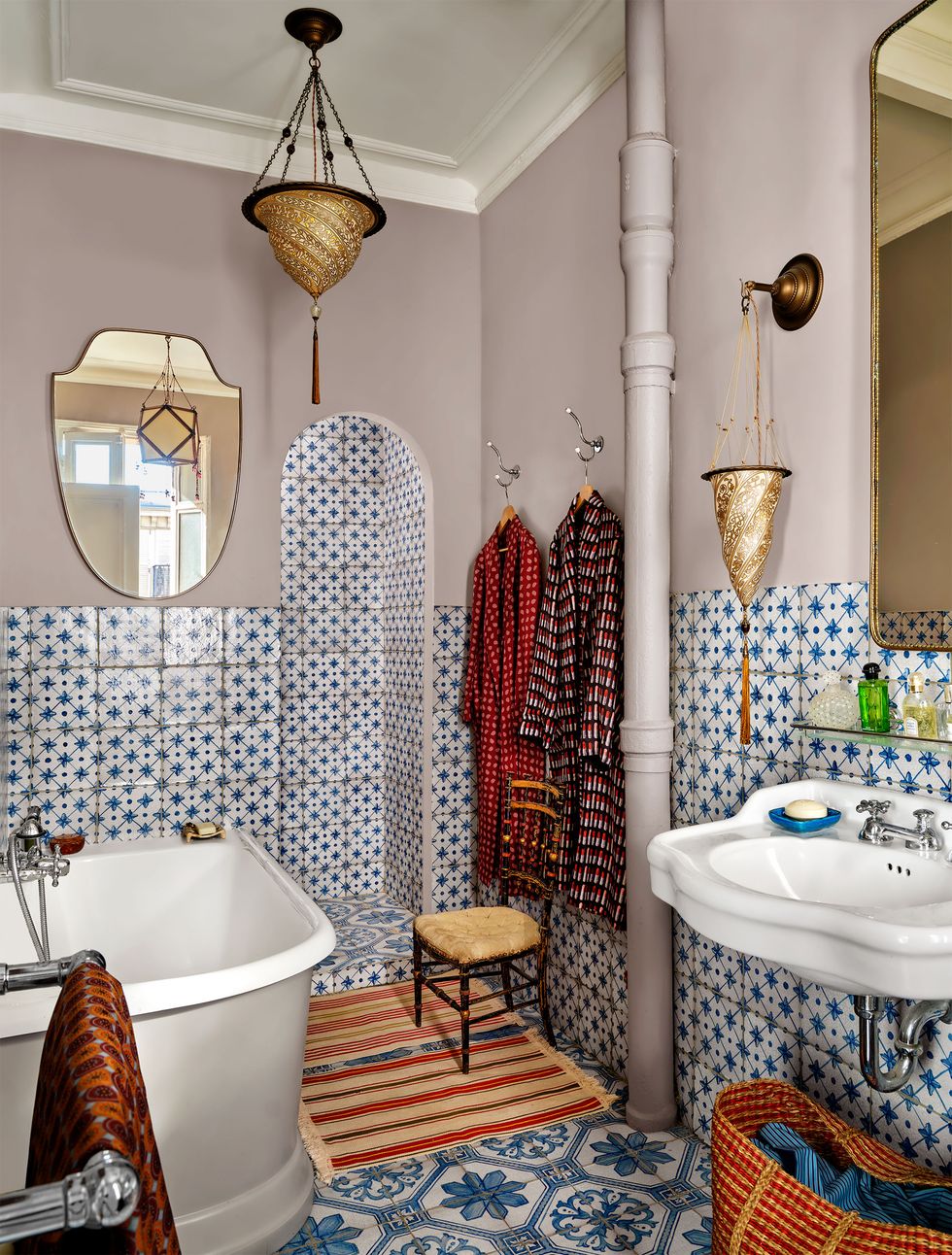 a bathroom has handmade blue and white tiles on the floor, halfway up the walls, and in the shower, matching cone shaped sconce and pendant, a deep bathtub, striped bath rug and two hanging bathrobes