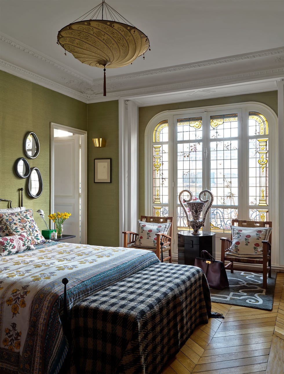 Marc Valeanu - ELLE DECOR an iron bed has flowered bedding and a checkered blanket, sage green walls, ornately patterned glass in a curved window frame, a decorative pendant with a hanging tassel, two wood armchairs with pillows