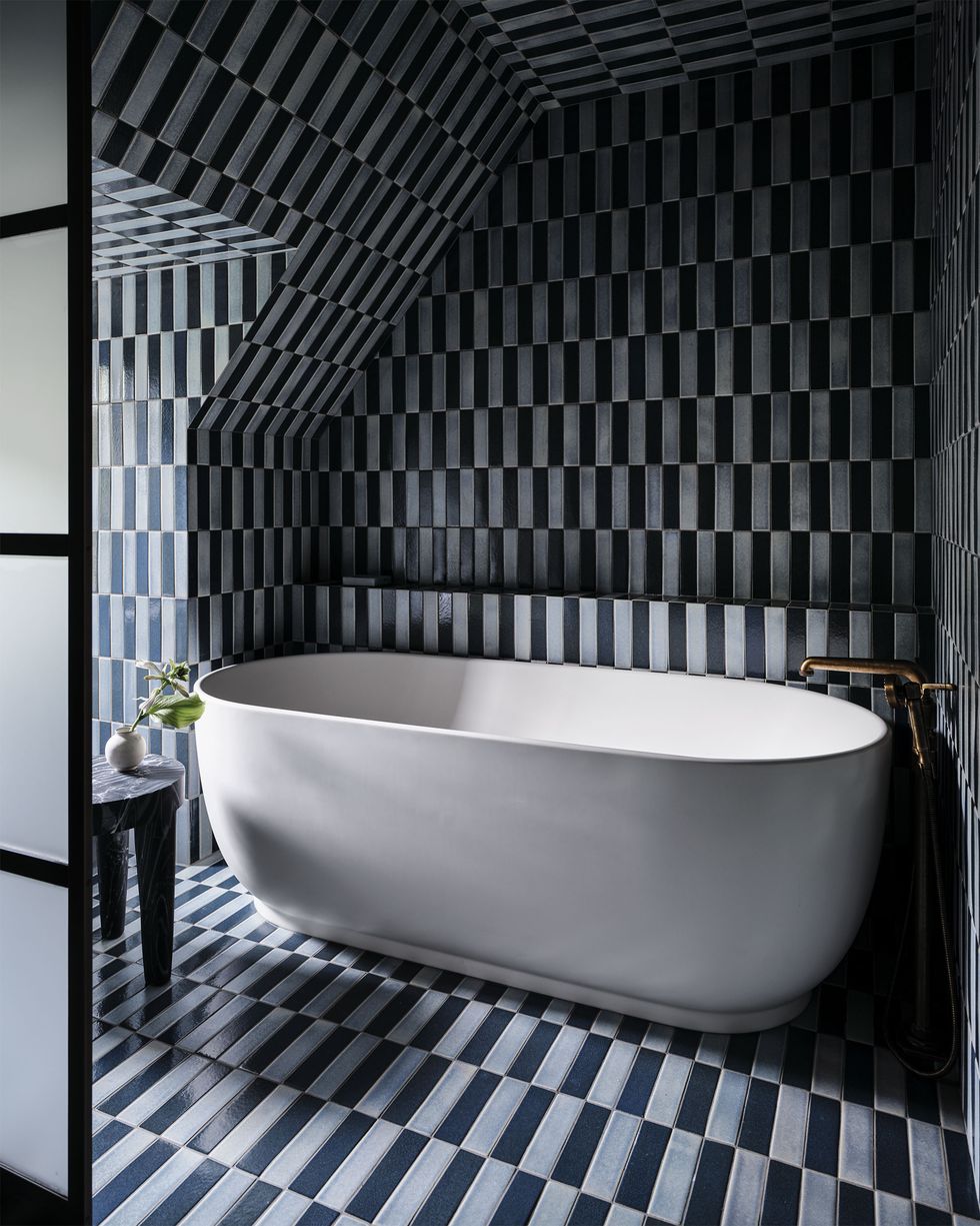 black and white rectangular tiles cover the bathroom walls, floor, and ceiling, a deep white tub is set in a nook, a three legged stool sits next to a frosted glass door