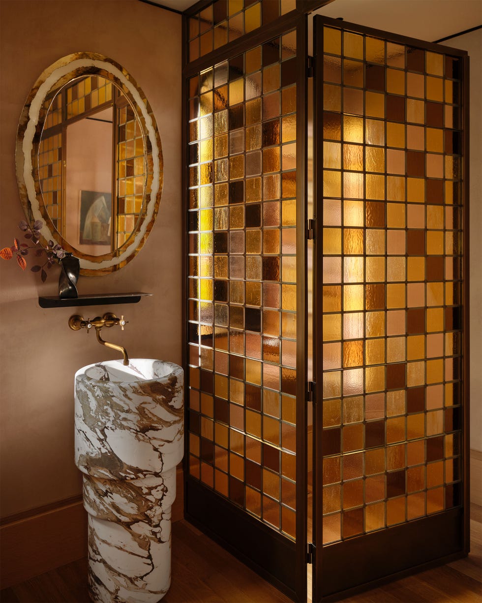 in a bathroom with brown walls, a wall with door is made of square glass tiles in shades of brown and amber, oval mirror above a marble pedestal sink with bronze fittings