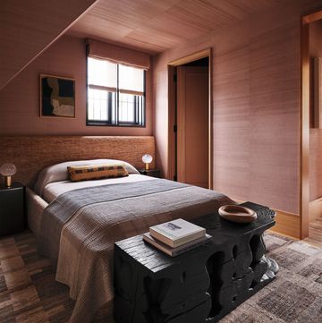 a bedroom has a rose colored paper on the walls and ceiling, a bed has a fabric headboard and chenille blankets with nightstands below a small window, a dark wood bench at foot of bed, and a patterned rug