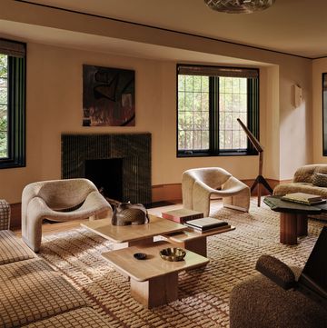 a living room is decorated in tones of brown and beige including two sofas, two modern armchairs, three travertine cocktail tables of different heights, a fireplace, floor lamp, and patterned rug