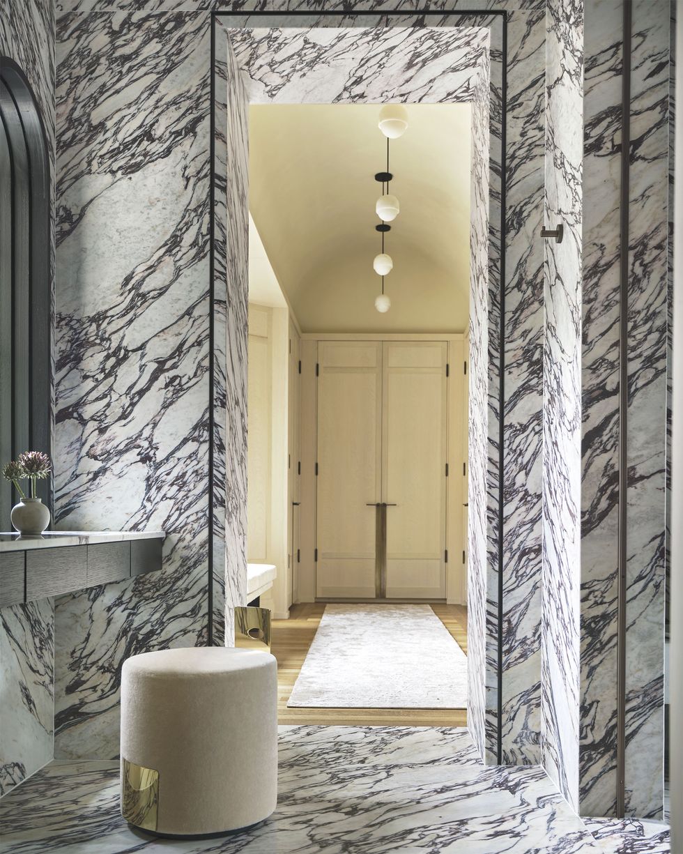 a bathroom in encased in a white marble with dark veins on the floor, a round stool sits in front of a vanity, and a door leads into a dressing room with a window seat and closets, small white globe pendants