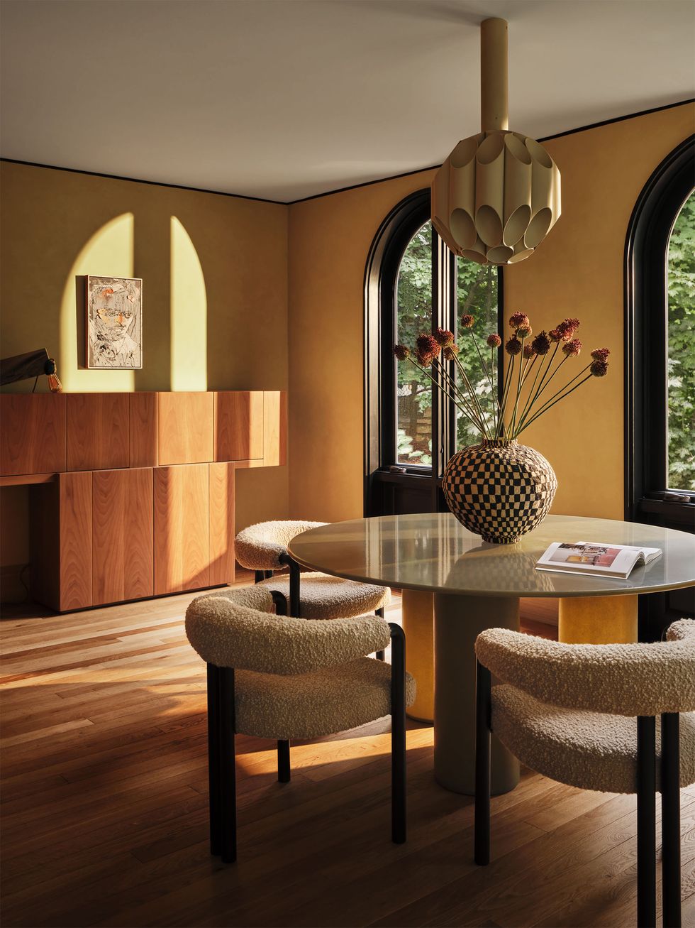 in a bar and lounge area is a wood floor, arched windows, a walnut sideboard, a round table with three boucle covered low back chairs, a multifaceted round pendant, and a checkerboard patterned vase