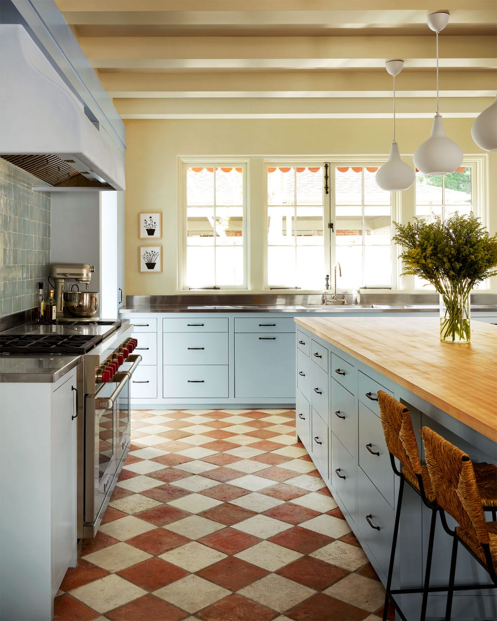 Ashe Leandro - ELLE DECOR kitchen with light blue cabinetry, light yellow walls, island with butcher block top and two rattan backed stools and three teardrop pendants, windows over sink, terra cotta and white diamond shape floor tiles
