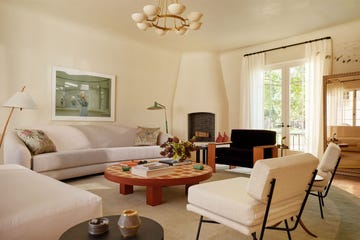 white living room with a curved light sofa, two floor lamps, round cocktail table, side tables, white slipper chair, two cushioned chairs with metal base, black chair, adobe fireplace in corner, window with sheers