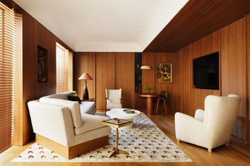 living room with wood paneled walls, a recessed white ceiling, built in tv screen, two white boucle chairs and a curved sofa, small built in desk with chair and sconce above, geometric rug, wood blinds