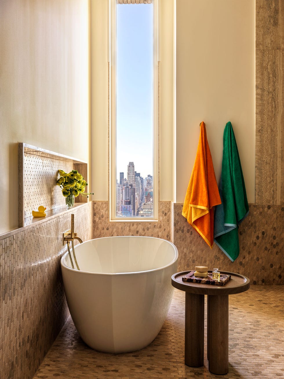 a bathroom has a window with cityscape view, deep white oval bathtub, round wooden table, floor and halfway up walls are small mosaic tiles in shades of brown, a red and a green towel hang on hooks