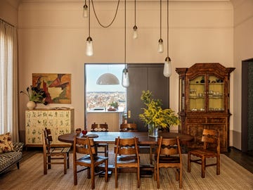 dining room with oblong wood table and eight vintage chairs, entry door from kitchen flanked by a flowered cabinet and a glass fronted cabinet, chandelier with long teardrop lights, small settee by window, neutral rug