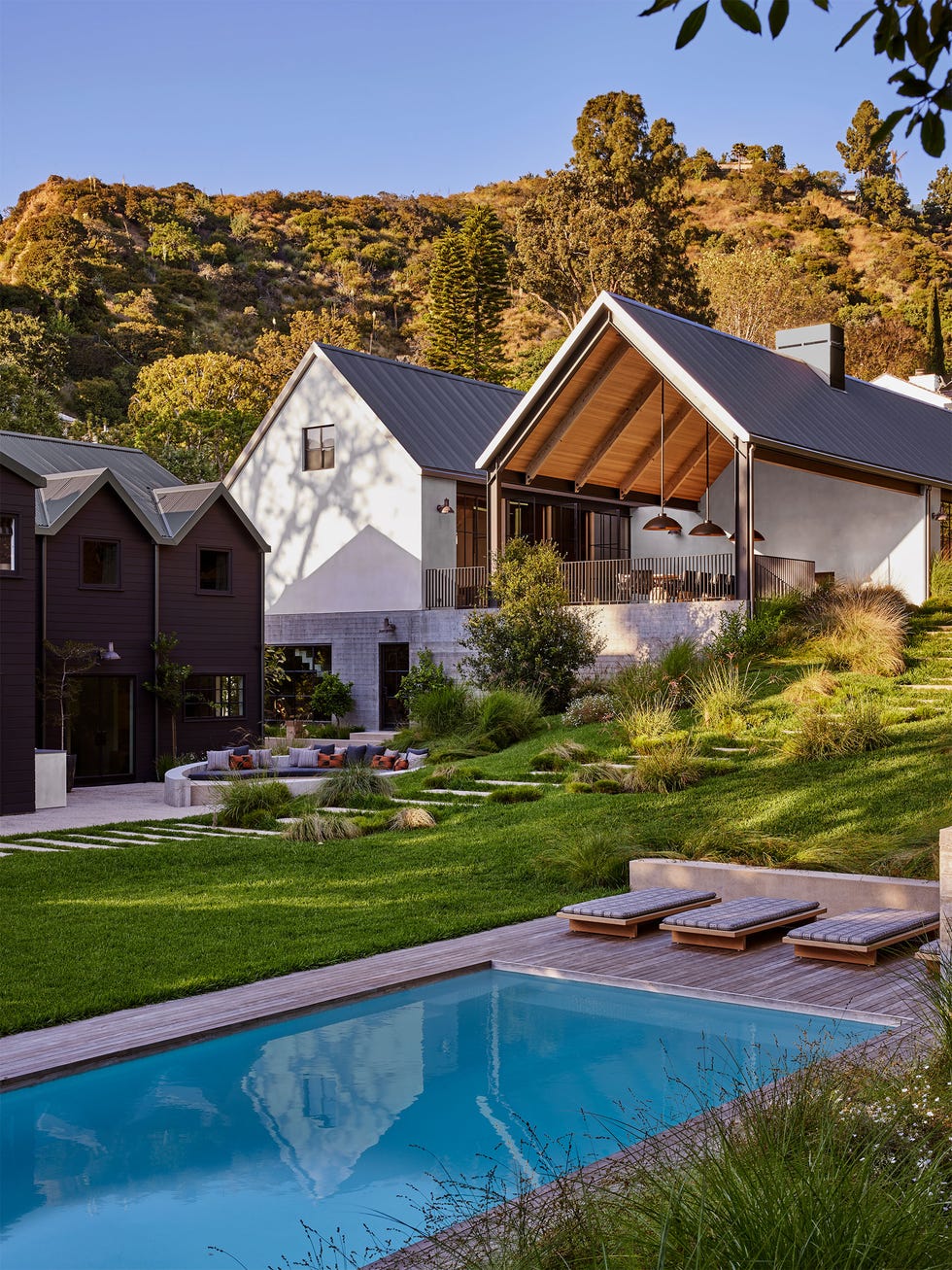 rectangular pool with chaises, dark exterior house with gabled roof, outdoor seating area, sloping hill with bushes and grasses, concrete steps to a modern house with pitched roofs, open terrace with pendants