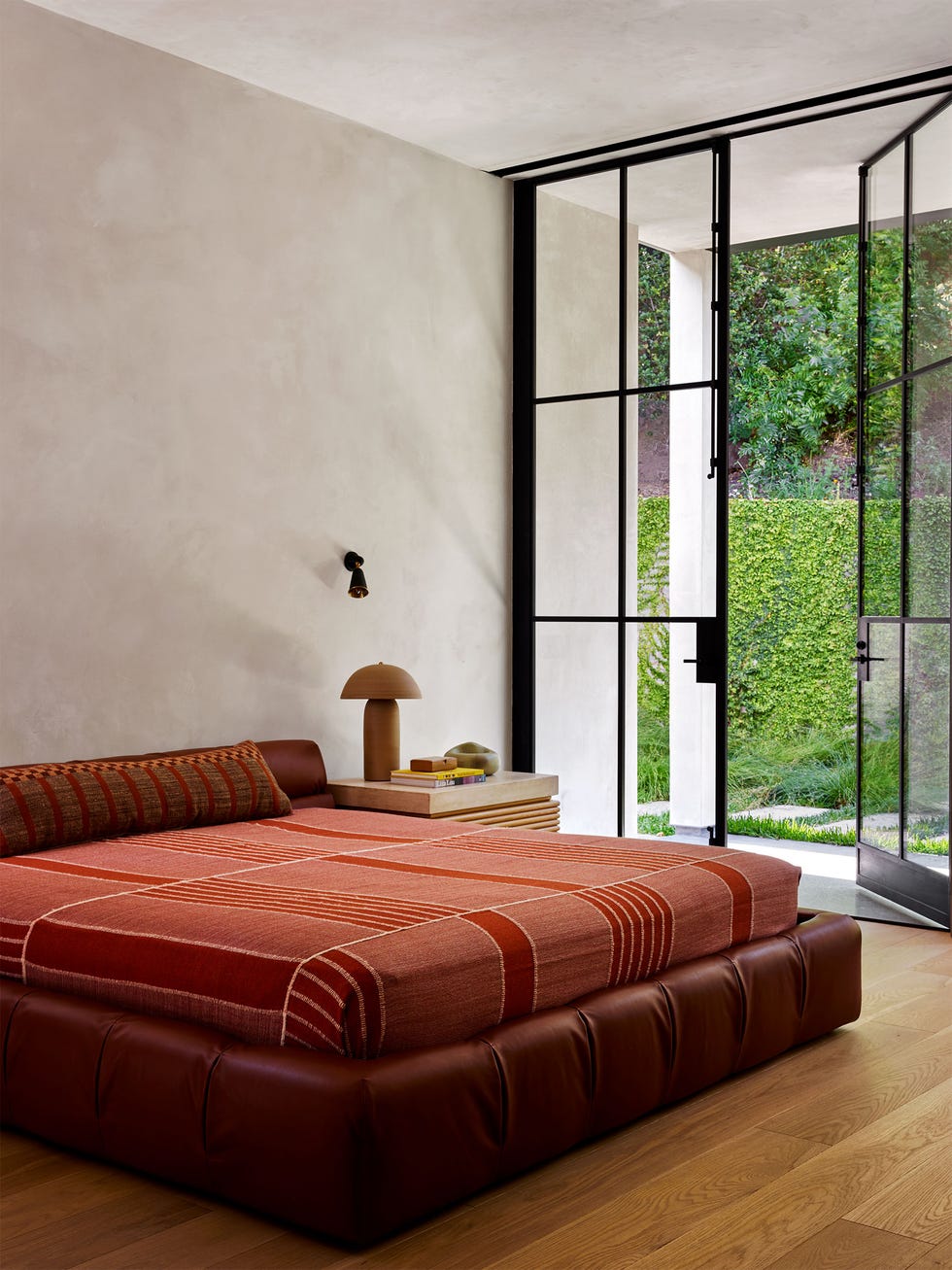 bedroom with plaster walls and one wall of square paned windows with a door leading outside, cognac leather frame bed, striped reddish bedcover, sconce above nightstand with lamp and books, wood floor