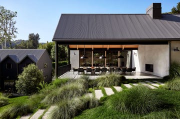 two houses, one with gabled roof, the other with a slanted roof over a large dining table with 18 chairs, three pendants on a deck, two open walls, one concrete, the other glass, concrete steps, bushes and grasses