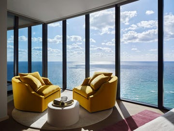 living room with floor to ceiling windows looking out on the turquoise ocean with white clouds in the sky, two mustard colored swivel chairs on a round rug set on a larger rug, round white cocktail table