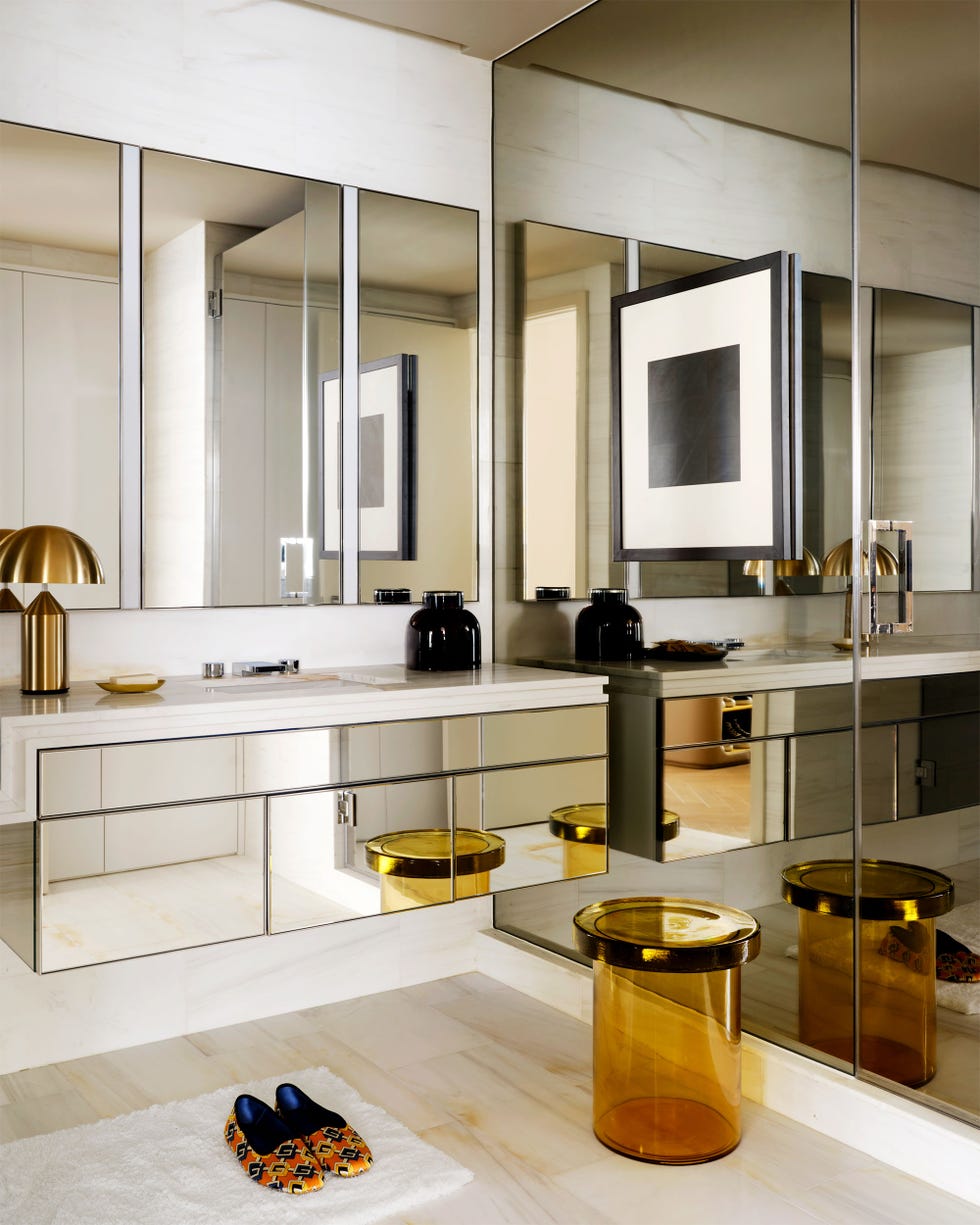 a bathroom with white walls and light floor, counter with sink and a lamp has a mirrored front cabinets, three mirrors on wall above countertop, amber glass stool, bright silk slippers on a small white bathmat