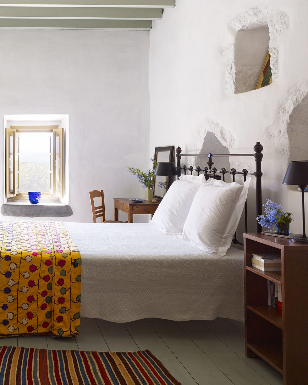 Patmos Greece - ELLE DECOR airy white metal bed in a room with white plank floors and double door and window at far end and a wooden table and chair and