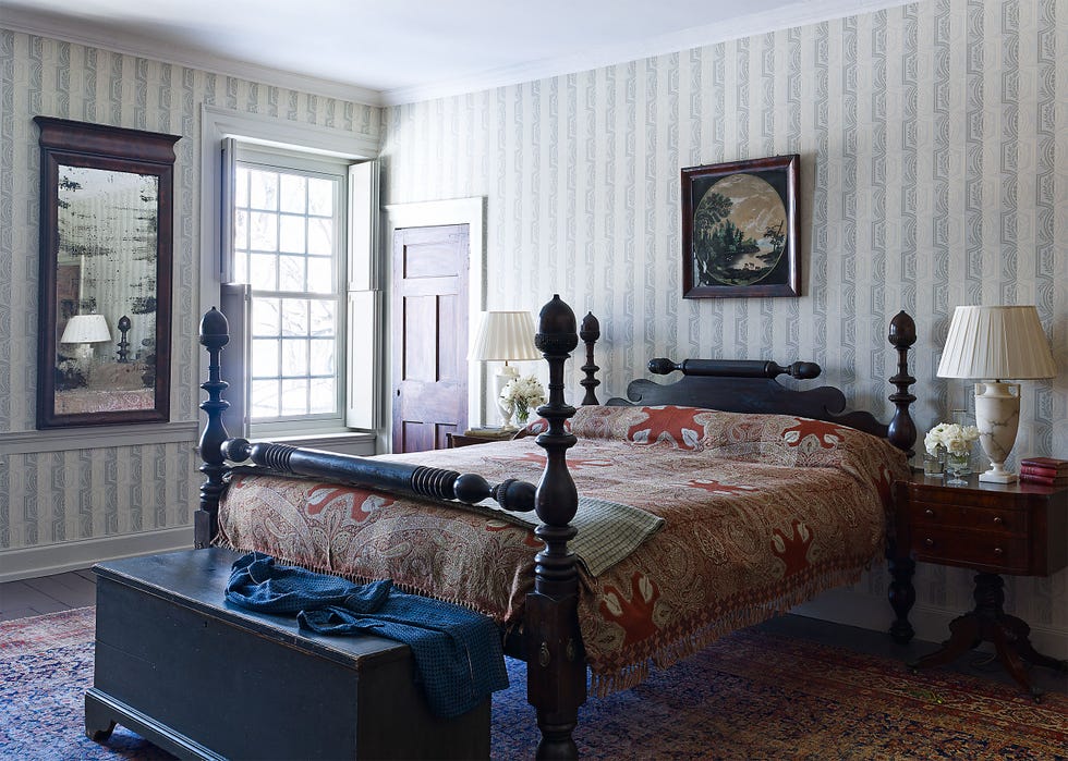 bedroom with white and light gray striped walls and and bed frame with four heavy turned posts and a white lamp on a nightstand and a heavy chest at the foot