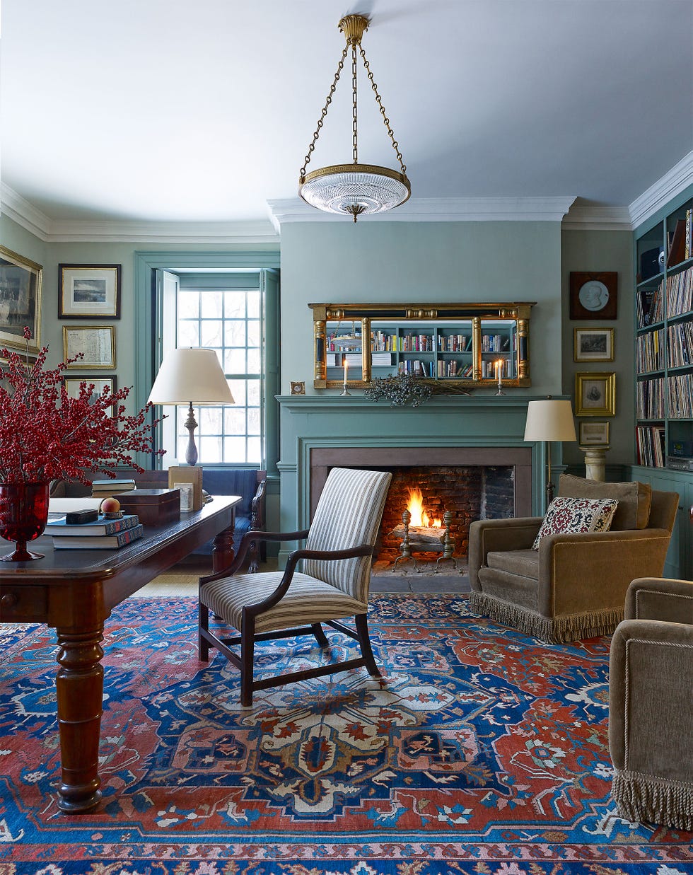 library room with persian rugs and an elegant table desk and chair and a blue wall with lit fireplace and a long gift mirror over it that reflects the shelves on the opposite wall and inset shelving along the sides
