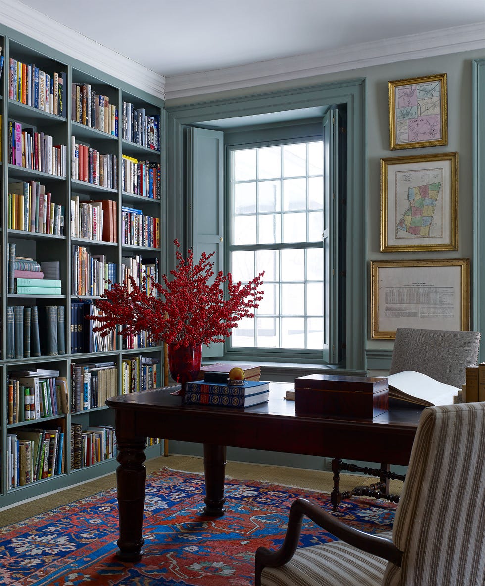 the room features a large bookshelf filled with a variety of books, while a wooden desk sits at the center with a few open books, a decorative box, and a fruit a striking bouquet of red flowers in a glass vase adds a pop of color to the side, an armchair with a striped pattern provides seating, and the room is illuminated by natural light streaming through a window
