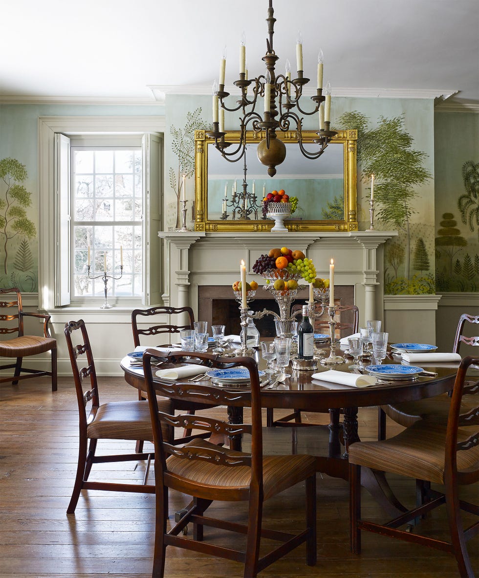 a spacious dining room featuring a large round wooden table surrounded by wooden chairs the table is dressed with fine china, crystal glassware, and neatly arranged utensils, creating an inviting atmosphere a centerpiece of fresh fruit in a glass bowl is complemented by two candlesticks with lit candles the walls are adorned with a scenic wallpaper depicting trees and foliage natural light streams in through large windows, and a chandelier hangs from the ceiling, adding a warm ambiance to the room