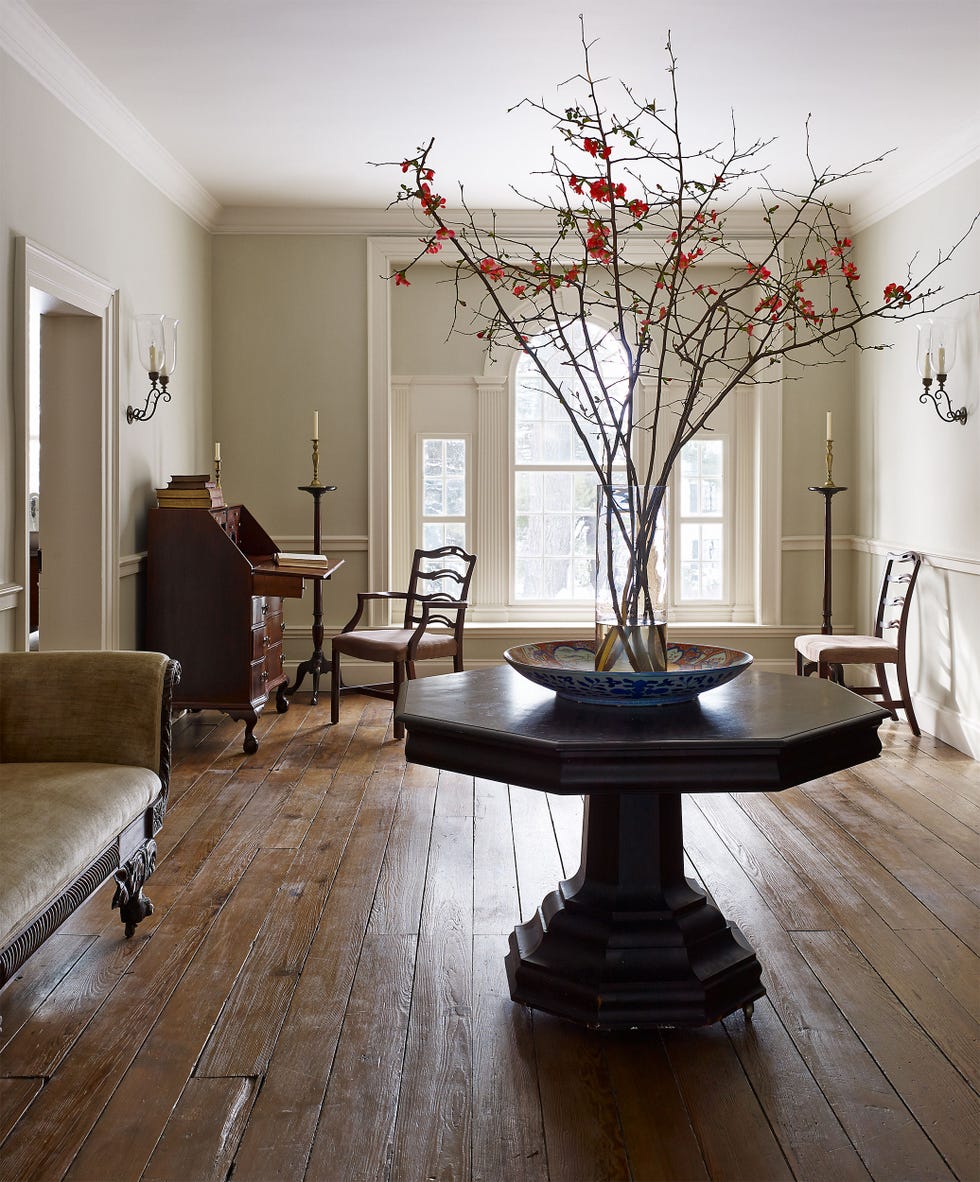 a spacious room featuring wooden flooring and elegant furnishings the focal point is an octagonal table adorned with a colorful decorative bowl, which holds branches with red flowers around the room, there are a couple of chairs, a writing desk, and candle sconces mounted on the walls natural light streams through large windows, enhancing the warm atmosphere